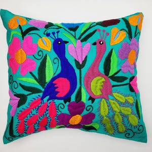 Könnte beinhalten: Türkisfarbenes, quadratisches Kissen mit gesticktem Design von zwei stilisierten Pfauen, einem blauen und einem rosa, umgeben von bunten Blumen und Blättern. Das Kissen ist mit leuchtenden Farben wie Pink, Orange, Lila und Grün verziert.