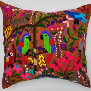 Könnte beinhalten: Ein braunes Kissen mit bunten gestickten Vögeln, Blumen und Blättern. Das Kissen zeigt einen Papagei, einen Blauvogel, einen Schmetterling und andere florale Designs.