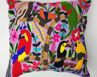May include: Embroidered decorative pillow with a vibrant design featuring colorful birds, flowers, and foliage on a lavender background. The pillow showcases a tree with various birds, including a parrot, toucan, and other species.