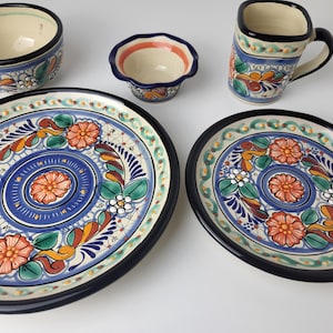 Könnte beinhalten: Eine Sammlung handbemalter Keramikgeschirr mit floralen Designs. Enthält eine große ovale Platte, einen kleineren runden Teller, eine Schüssel, eine kleine Schale und einen Becher. Die Teile haben einen cremefarbenen Hintergrund mit blauen, orangefarbenen, grünen und schwarzen Akzenten.