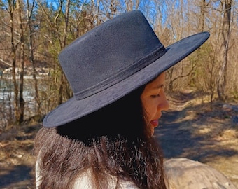 May include: A solid black wide-brimmed hat with a flat crown and a matching black band around the base. The hat features a textured, suede-like finish and a structured silhouette, providing a clean and uniform appearance across the entire surface.
