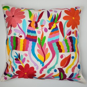 Puede incluir: Cojín cuadrado blanco con bordado colorido. El diseño presenta dos animales estilizados con cuerpos a rayas arcoíris y motivos florales y de hojas en rosa, naranja, rojo y verde. Un elemento decorativo para el hogar.