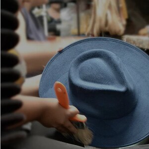 May include: A person is brushing a dark blue felt fedora hat with an orange-handled brush. The hat is sitting on a surface.