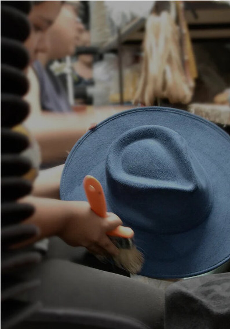 May include: A person is brushing a dark blue felt fedora hat with an orange-handled brush. The hat is sitting on a surface.