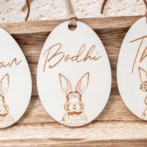 May include: Three wooden Easter egg ornaments with bunny illustrations and personalized names. The names are Bodhi, Liam, and Thea. The ornaments are hanging from brown leather cords.