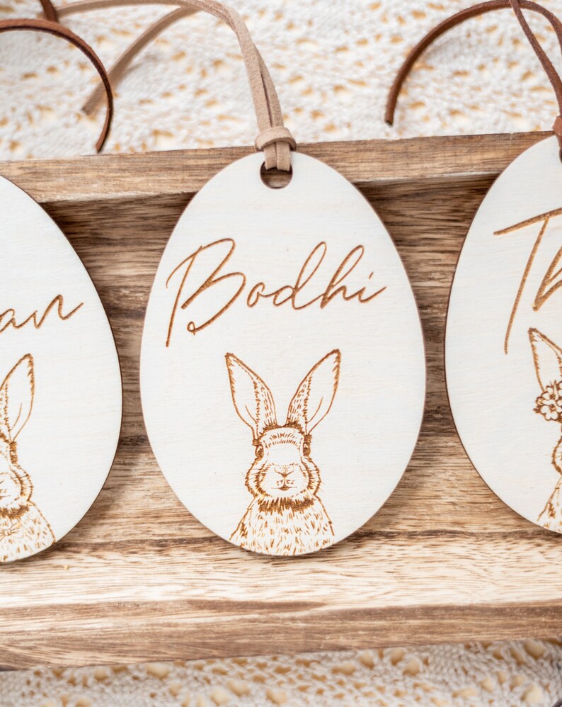 May include: Three wooden Easter egg ornaments with bunny illustrations and personalized names. The names are Bodhi, Liam, and Thea. The ornaments are hanging from brown leather cords.