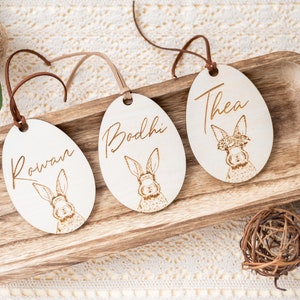 May include: Three wooden Easter egg ornaments with bunny illustrations and personalized names: Rowan, Bodhi, and Thea. The ornaments are hanging from brown twine and are displayed on a wooden tray.