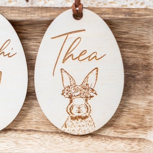 May include: A wooden Easter egg ornament with a bunny wearing a flower crown. The egg is engraved with the name "Thea".