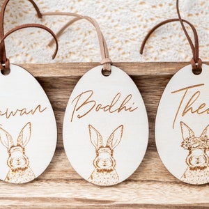 May include: Three wooden Easter egg ornaments with bunny illustrations and personalized names: Rowan, Bodhi, and Thea. Each egg has a brown leather cord for hanging.