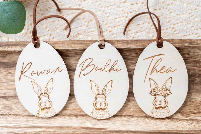 May include: Three wooden Easter egg ornaments with bunny illustrations and personalized names: Rowan, Bodhi, and Thea. Each egg has a brown leather cord for hanging.