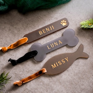 May include: Three personalized pet name tags in the shapes of a bone, a fish, and a rectangle with a paw print. The tags are made of brown and gray acrylic with gold lettering. The names on the tags are Benji, Luna, and Missy.