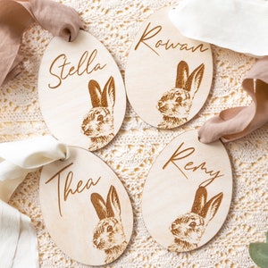 May include: Four wooden Easter egg ornaments with bunny illustrations and personalized names: Stella, Rowan, Thea, and Remy. Each egg is tied with a ribbon.
