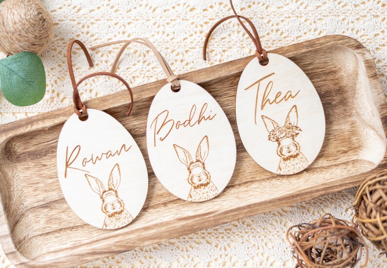 May include: Three wooden Easter egg ornaments with bunny illustrations and personalized names: Rowan, Bodhi, and Thea.