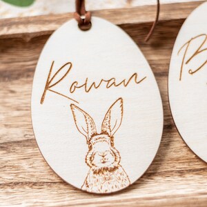 May include: A white wooden egg-shaped ornament with a brown leather cord. The ornament is engraved with the name "Rowan" and a drawing of a rabbit.