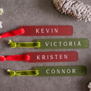 May include: Four personalized gift tags with red and green velvet ribbon. The tags are made of clear acrylic and have the names Kevin, Victoria, Kristen, and Connor printed in white.