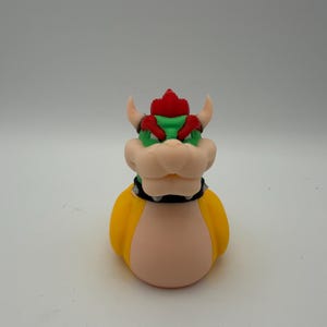May include: A rubber duck designed to resemble the character Bowser, featuring a yellow body, a peach-colored chest and snout, green skin on the head, a red mohawk, two small horns, and a black spiked collar around the neck.