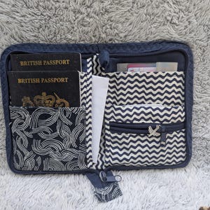 Op de afbeelding: Marineblauwe reisportemonnee met meerdere compartimenten, waaronder sleuven voor paspoorten en kaarten. De binnenkant heeft een wit en marineblauw zigzagpatroon en een marineblauw wervelontwerp. Twee Britse paspoorten zijn zichtbaar.