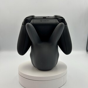 Op de afbeelding: Een zwarte Xbox-controller rust op een zwarte konijnvormige standaard. De standaard staat op een wit draaiend platform. De Xbox-controller is zwart met het Xbox-logo zichtbaar. De konijnenstandaard heeft lange oren en een gladde afwerking.