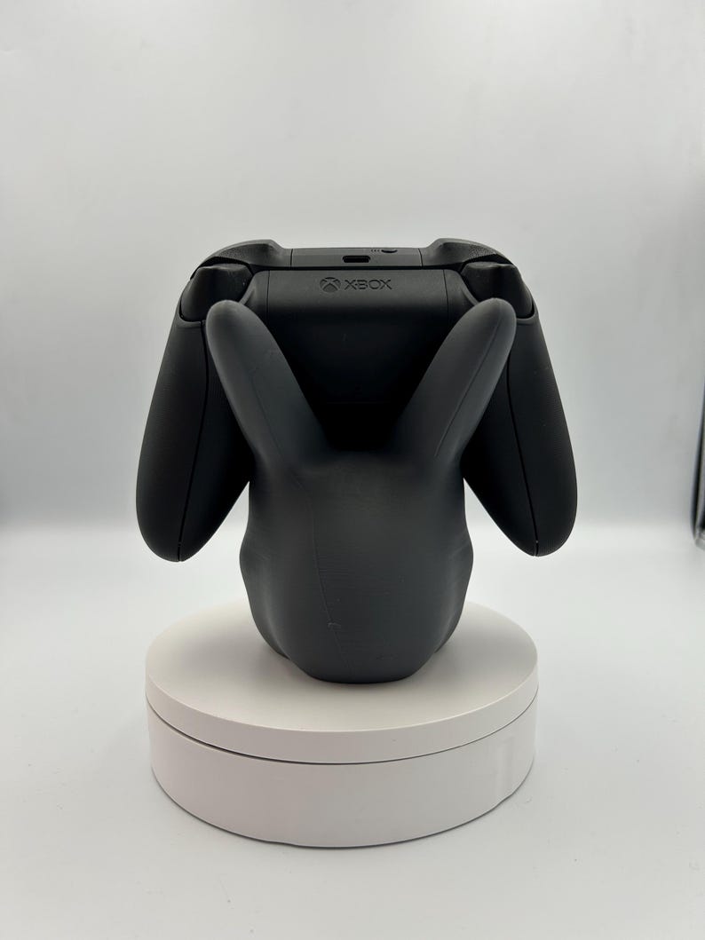 Op de afbeelding: Een zwarte Xbox-controller rust op een zwarte konijnvormige standaard. De standaard staat op een wit draaiend platform. De Xbox-controller is zwart met het Xbox-logo zichtbaar. De konijnenstandaard heeft lange oren en een gladde afwerking.