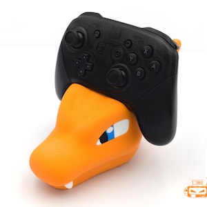 Op de afbeelding: Een oranje en witte Charizard-thema controllerstandaard met een zwarte Nintendo Switch Pro controller. De standaard heeft een gedetailleerd ontwerp van de kop van het Pok&eacute;mon-personage, met een blauw oog en witte accenten.