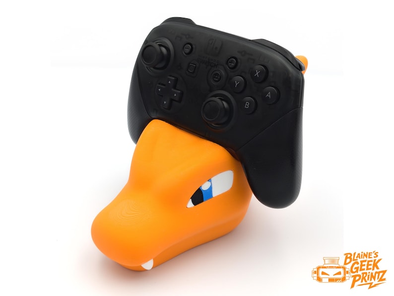 Op de afbeelding: Een oranje en witte Charizard-thema controllerstandaard met een zwarte Nintendo Switch Pro controller. De standaard heeft een gedetailleerd ontwerp van de kop van het Pok&eacute;mon-personage, met een blauw oog en witte accenten.