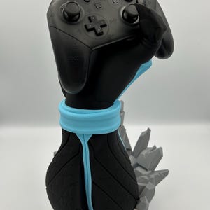 Może przedstawiać: Czarny i niebieski uchwyt na kontroler do kontrolera Nintendo Switch Pro. Uchwyt ma kształt dłoni, z jasnoniebieską opaską na nadgarstek i skalistą podstawą. Kontroler jest czarny z szarymi przyciskami.