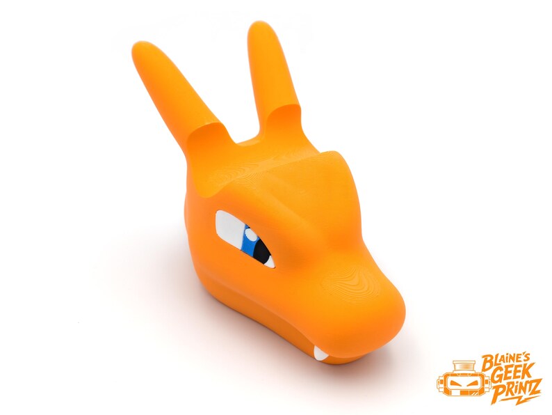 Op de afbeelding: Oranje 3D-geprinte Charizard telefoonhouder. De houder heeft de vorm van Charizards hoofd, met twee rechtopstaande hoorns, witte tanden en blauwe en witte ogen. Het product staat op een witte achtergrond. Het logo "Blaine's Geek Printz" staat rechtsonder.