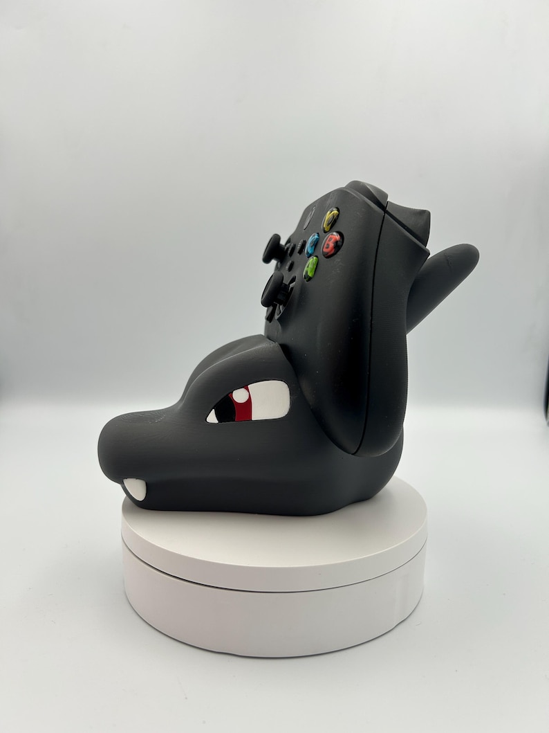 Op de afbeelding: Zwarte controllerhouder in de vorm van een draak. De draak heeft een wit oog met een rode pupil en een witte tand. De controllerknoppen zijn zichtbaar op de bovenkant van de drakenkop. De houder staat op een witte draaiende standaard.