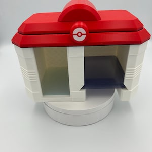 May include: A 3D-printed white and red storage box with a red roof and a white base. The box has a circular emblem on the front and two open compartments. The box is designed to resemble a building.