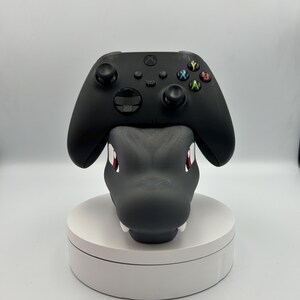 Op de afbeelding: Een zwarte Xbox-controller rust op een grijze drakenkopvormige standaard. De draak heeft rode ogen en witte tanden. De standaard staat op een witte, ronde basis, voor gaming.