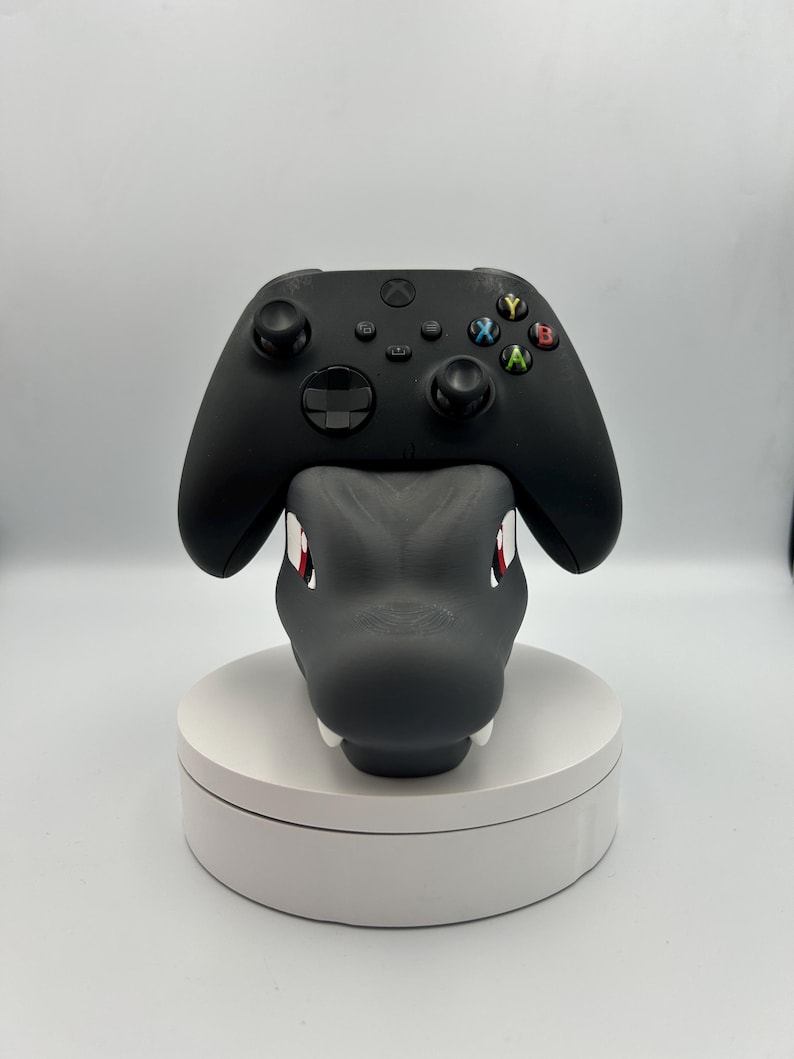 Op de afbeelding: Een zwarte Xbox-controller rust op een grijze drakenkopvormige standaard. De draak heeft rode ogen en witte tanden. De standaard staat op een witte, ronde basis, voor gaming.