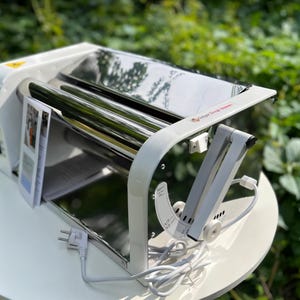 May include: A white and stainless steel dough roller machine featuring a top-mounted reflective metal cylinder, a side-mounted adjustable lever with measurement markings, a power switch, and an attached white electrical cord with a plug.