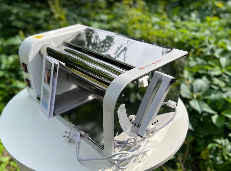 May include: A white and stainless steel dough roller machine featuring a top-mounted reflective metal cylinder, a side-mounted adjustable lever with measurement markings, a power switch, and an attached white electrical cord with a plug.