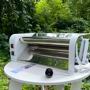 May include: A white electric dough roller machine featuring two horizontal stainless steel rollers, a white plastic frame, and two red power switches on the side. A white power cord with a black plug extends from the side of the unit.