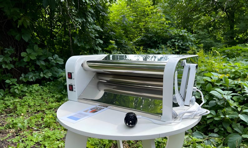 May include: A white electric dough roller machine featuring two horizontal stainless steel rollers, a white plastic frame, and two red power switches on the side. A white power cord with a black plug extends from the side of the unit.