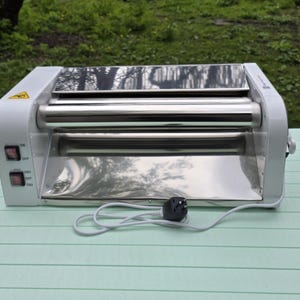 May include: A white and silver laminator machine with two red on/off switches. The machine has a reflective stainless steel surface and rollers. A black plug and white cord are connected to the machine.