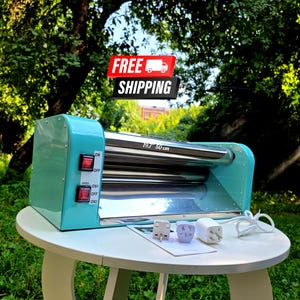 May include: A turquoise dough roller machine with a 19.7 inch width, featuring two red power switches on the side panel, a polished metal roller assembly, and three white electrical power adapters with a white cord resting on the table surface.