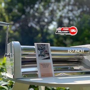 May include: A manual dough sheeter for home use, with a 19.7-inch roller. The machine is silver and white, with a crank handle. A small instruction manual is visible. The background is a natural green.