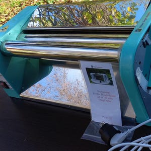 May include: A teal and silver electric dough sheeter with two polished metal rollers. A white information card is attached, reading: Professional Electric Dough sheeter 220 or 110 volts Dough roller. A white power cord with a plug is visible at the base.