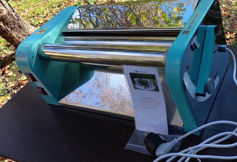 May include: A teal and silver electric dough sheeter with two polished metal rollers. A white information card is attached, reading: Professional Electric Dough sheeter 220 or 110 volts Dough roller. A white power cord with a plug is visible at the base.