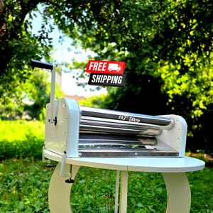 May include: A white manual rolling machine with two horizontal stainless steel rollers. The machine features a black handle on the left side and a metal clamp for table mounting. The text 19.7" 50cm is printed on the upper roller surface.