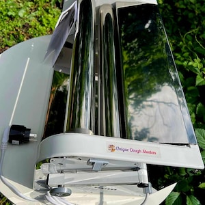 May include: A white electric dough sheeter featuring two polished stainless steel rollers, a white base, and a power cord with a plug. A label on the front reads Unique Dough Sheeters, and a yellow warning triangle sticker is on the top housing.