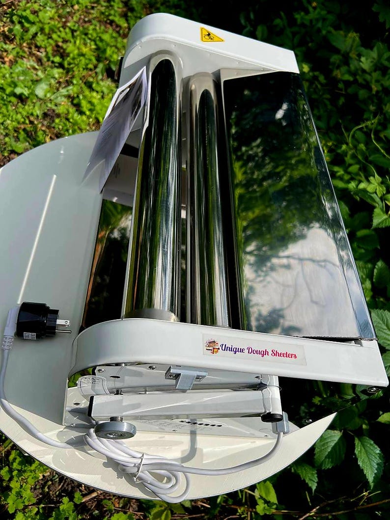 May include: A white electric dough sheeter featuring two polished stainless steel rollers, a white base, and a power cord with a plug. A label on the front reads Unique Dough Sheeters, and a yellow warning triangle sticker is on the top housing.