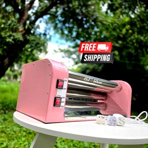 Può includere: Una macchina rotante per tazze di colore rosa con due rulli metallici orizzontali, un pannello di controllo laterale con due interruttori rossi e un cavo di alimentazione bianco. Il dispositivo riporta un'etichetta di 50cm di lunghezza.