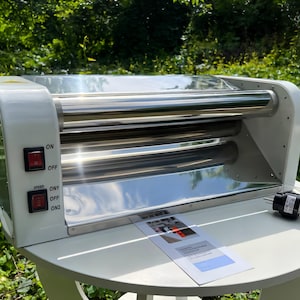 May include: A white electric hot dog roller machine with stainless steel rollers. The side panel features two red power switches labeled ON, OFF, and ON1, ON2. A power cord with a plug extends from the side, and a small instruction manual rests in front.