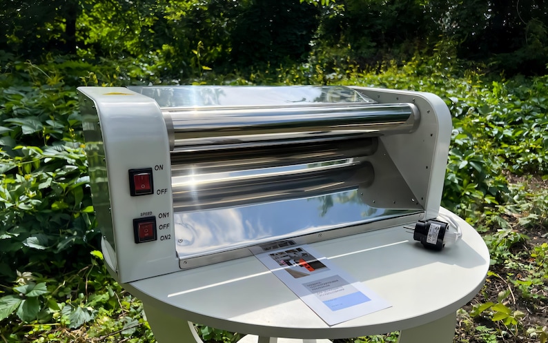 May include: A white electric hot dog roller machine with stainless steel rollers. The side panel features two red power switches labeled ON, OFF, and ON1, ON2. A power cord with a plug extends from the side, and a small instruction manual rests in front.