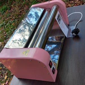 May include: A pink electric roller machine with two polished metal cylinders, a white power cord, and two black power switches on the side. A yellow warning triangle icon is visible on the pink frame, and a small instruction tag is attached.