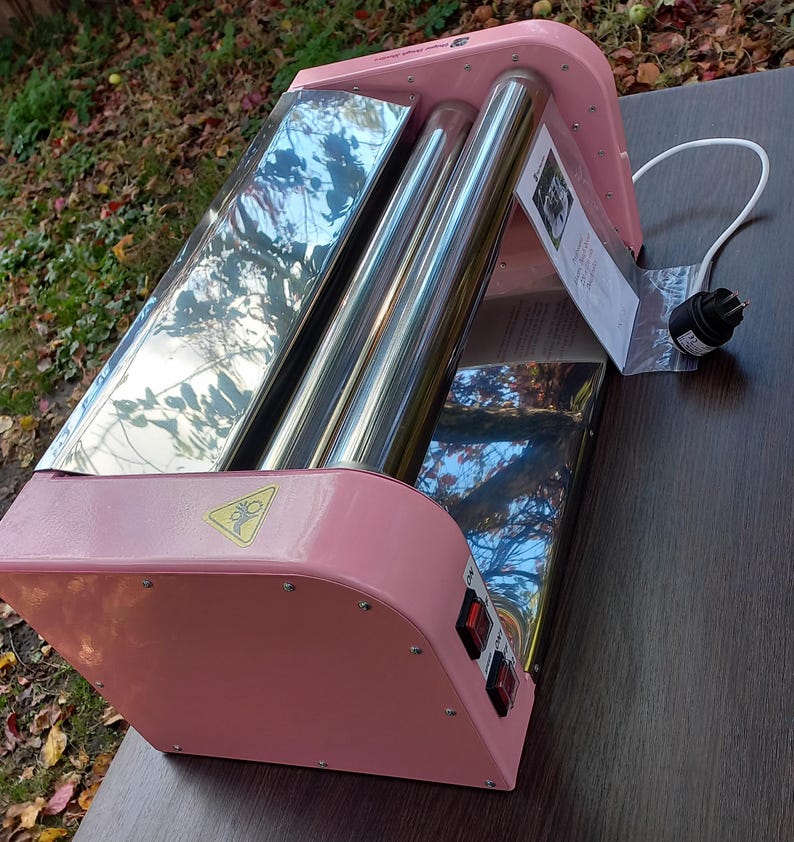 May include: A pink electric roller machine with two polished metal cylinders, a white power cord, and two black power switches on the side. A yellow warning triangle icon is visible on the pink frame, and a small instruction tag is attached.