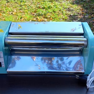 May include: A light blue electric dough roller machine with a polished chrome finish on the central rollers and a flat metal surface. The device features two red power and speed switches on the left side, a yellow warning label, and a white power cord.
