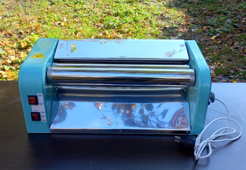 May include: A light blue electric dough roller machine with a polished chrome finish on the central rollers and a flat metal surface. The device features two red power and speed switches on the left side, a yellow warning label, and a white power cord.